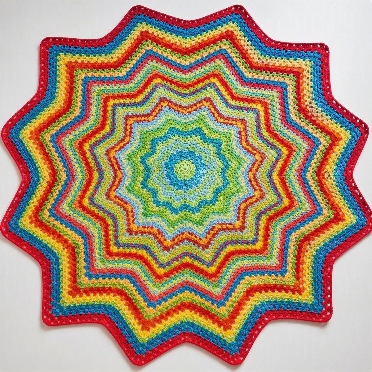 Twelve-point star-shaped crochet blanket in concentric rainbow rings, from a teal centre out to a red scalloped edge.