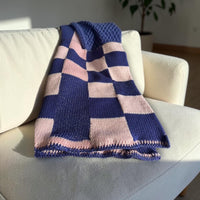 Navy and pale pink checkerboard knit blanket folded over a cream armchair, with a textured panel and contrast crochet edge.